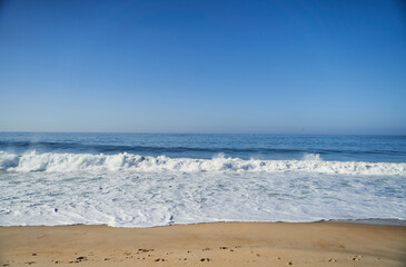 A closeup shot of the ocean with wind waves crashing onto the shore, creating a beautiful pattern of furrows in the water. The landscape is enhanced by cumulus clouds in the sky