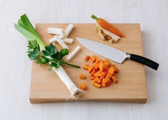 Fresh vegetables including carrots, celery, and leek are chopped on a wooden cutting board. A sharp knife rests beside them, ready for meal preparation in the kitchen.