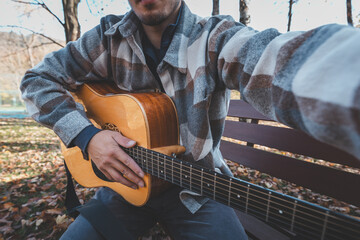 Man playing acoustic guitar outdoors in autumn, wearing a plaid shirt, sitting on a wooden bench...