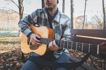 Man playing acoustic guitar outdoors in autumn, wearing a plaid shirt, sitting on a wooden bench...