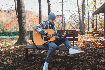 Young man sitting on a bench in a scenic autumn park, playing an acoustic guitar, surrounded by colorful trees and fallen leaves. Capturing calmness, creativity, and nature