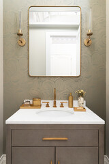 A bathroom sink detail with a gold faucet and decorations, a patterned wallpaper, white marble countertop, and a snake skin cabinet.