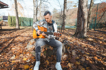 Focused guitarist in a plaid shirt strums an acoustic guitar while sitting on a stone bench amidst a golden autumn landscape. Sunlight enhances the warm, peaceful setting