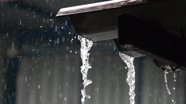 A rain gutter is dripping water. The water is falling from the gutter and splashing onto the ground. The scene is dark and moody, with the rain creating a sense of melancholy