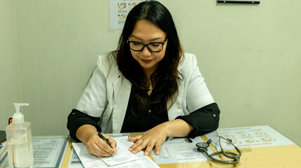A female doctor in a white coat writes notes at her desk. An eye chart and handwashing instructions are visible on the wall, emphasizing a clinical setting.
