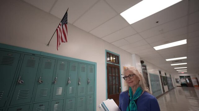 A middle-aged woman teacher walks through a school hallway holding a book. Brightly colored lockers line the walls under a hanging American flag.