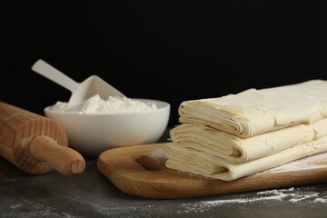 Board with raw puff pastry dough, rolling pin and flour on dark table against black background, closeup