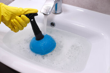 Clogged drain in bathroom sink. Hand in yellow gloves cleaning sink with plunger. Drain cleaning