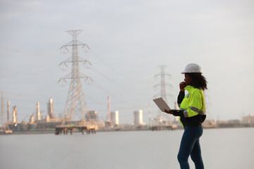 A Black female electrical engineer using a laptop computer to stand at the electricity station to see the electric power production plan at a high voltage electrode at the pier