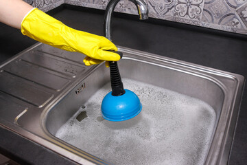 Hand in yellow gloves cleaning sink with plunger. Overflowing kitchen sink. Clogged drain.
