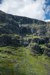 Rocky cliff mountain highland against a cloudy gray sky. Mountain sheep grazing in the grass on the cliff.