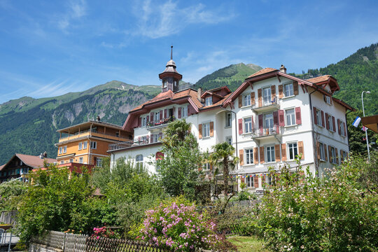 Charming architecture in Brienz, Switzerland, surrounded by lush greenery and vibrant flowers, with mountains in the background and a clear blue sky.