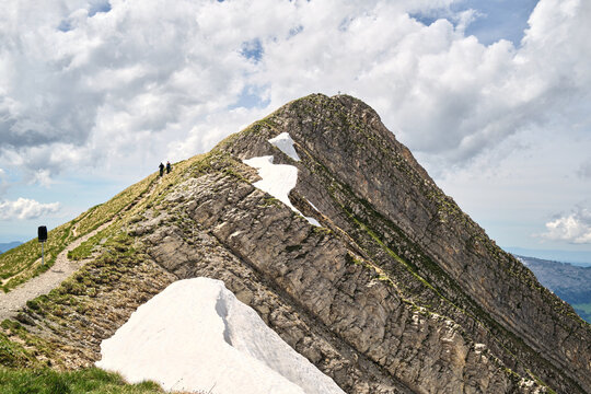 A breathtaking view of a mountainous landscape in Switzerland, showcasing steep rocky slopes and patches of snow, with hikers scaling the ridge near Brienz.