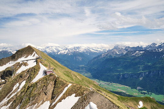 Stunning panoramic view of the Swiss Alps near Brienz, featuring snow-capped peaks, green valleys, and rocky ridges under a partly cloudy sky.