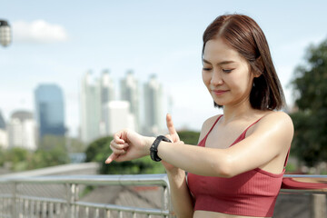 Fototapeta premium Short haired sporty girl in sportswear looks and touches smartwatch after jogging work out for exercise on city street. Happy smiling Asian woman do sport fitness outside. Healthy outdoor activity.