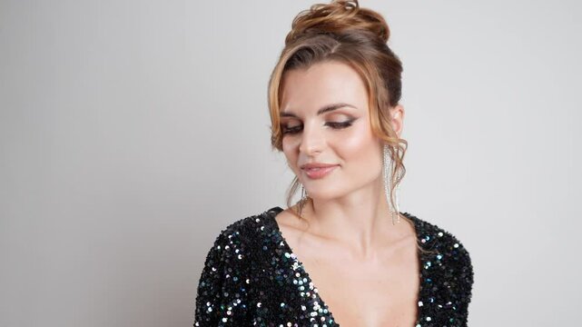 A beautiful woman with model looks in a low-cut, shimmering sequined dress with flawless evening makeup and an elegant updo poses with a shy smile for a studio portrait on a white background.