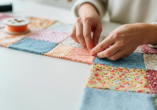 Close-up of hands using needle and thread, stitching together colorful fabric squares on white table, making patchwork quilt