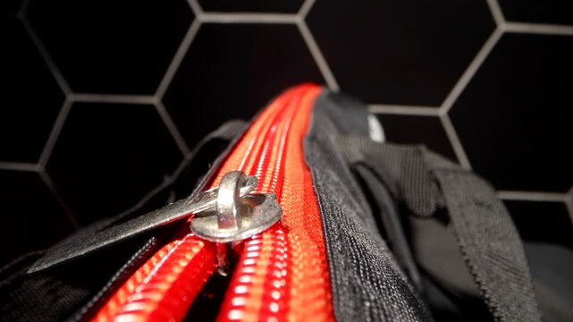 The contrast between the red zipper and the black fabric of the bag highlights the textures of the materials. The slider glitters against a minimalistic black background with a geometric pattern.