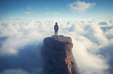 Person standing on a rocky peak surrounded by clouds at sunrise in a serene mountain landscape