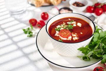Spanish Cold Tomato Gazpacho Soup garnished with parsley, feta cheese and spices with toasted bread slices, tomatoes and herbs on white table