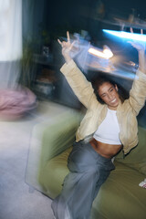Blurred motion of girl sitting on sofa with her arms raised and smiling during party at home