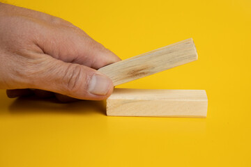 placing a wooden Jenga blocks on two blocks arranged neatly on a solid yellow background.