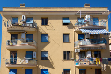 Yellow building with blue shutters and multiple balconies. Balconies have metal railings, awnings, and household items like laundry and chairs. Concept: Residential architecture.