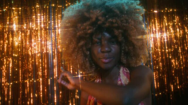 Close-up low key portrait of active and charismatic young African American lady with curly hair, wearing colorful open dress and shining necklace, dancing on tinsel background and having fun in club