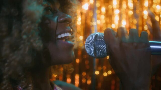 Side close-up of profile of young confident African American lady with curly hair singing into microphone and enjoying process on glittery tinsel background in club, nostalgic 70s