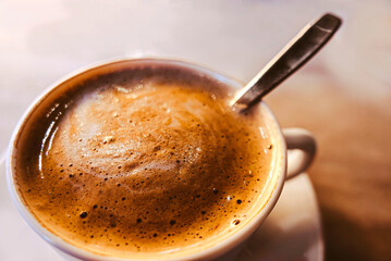 Close-up of a cup of frothy coffee with a spoon inside, on a wooden table, brown toned image