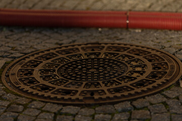 A circular manhole cover rests atop a cobblestone surface, surrounded by a prominently visible red pipe nearby