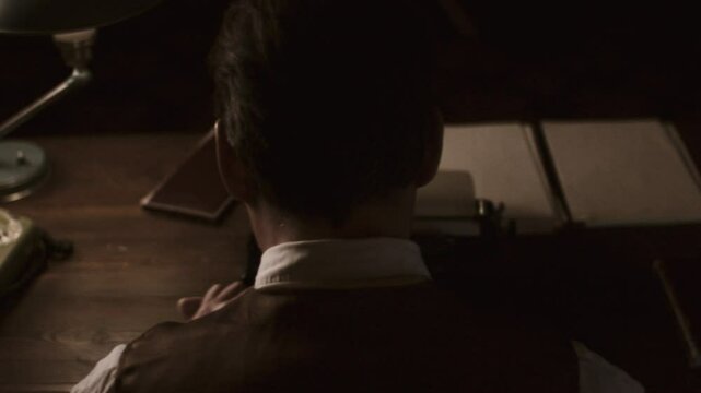 Vintage typewriter scene depicting young male author working intently, back turned, within shadowy office illuminated by warm desk lamp near aged telephone
