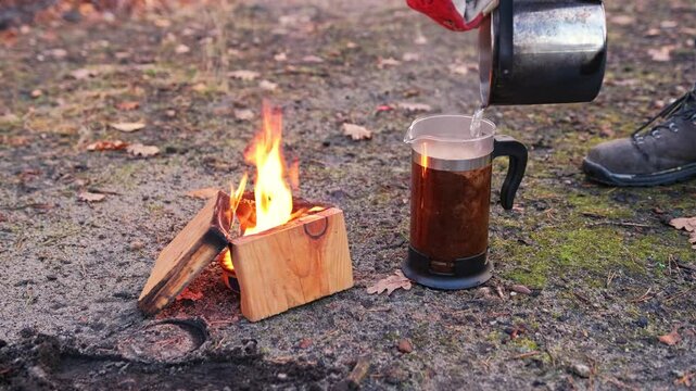 Male Hiker Tourist Pouring Boiling Hot Water into Ground Coffee Brewing French Press Jug at Improvised Camp Site with Buddy Burner Stove