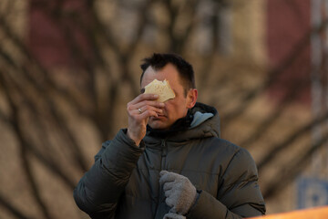 Hunger man stands outside in a bustling city, munching on food while dressed warmly to safeguard...