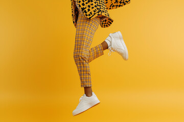 A persons legs in yellow plaid pants and white sneakers midjump against a vibrant yellow background. Minimalist fashion and dynamic movement.