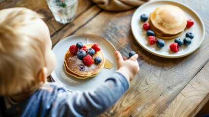 A toddler is reaching for a plate of pancakes adorned with vibrant berries on a warm wooden table. Fat Tuesday, National Pancake Day