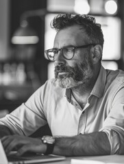Bearded man in office working on laptop, with focused gaze and glasses on.