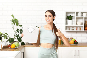 Sporty young woman with weight scales showing thumb-up in kitchen. Diet concept