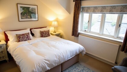  A cozy bedroom features a large bed with white linens, a nightstand with a lamp, and a window with a floral curtain, all set against a beige wall with a framed landscape painting.