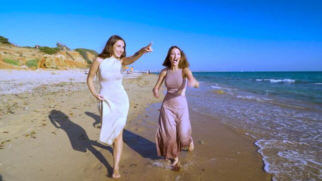 Dos chicas j&oacute;venes corriendo y sonriendo muy divertidas, por la orilla de la playa sobre la arena junto al mar, un magn&iacute;fico d&iacute;a soleado con cielo azul.