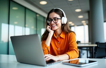 Young woman working on laptop inside modern office. Wears stylish orange shirt, headphones. Woman smiling, listening to online podcast audio course. Sits at workplace in contemporary office. Enjoying