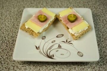 Delicate whole-grain bread bites topped with creamy spread, layered with cheese, ham, and a pickle slice secured with a toothpick, served on an elegant white floral-patterned plate.