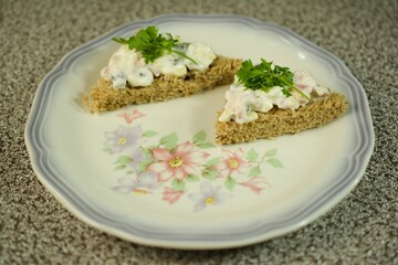 Tasty whole-grain sandwich triangles topped with a creamy mixture of fresh ingredients and decorated with parsley, served on a floral-patterned plate. Perfect for light snacks!