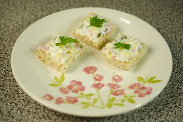 Three square mini sandwiches topped with creamy salad, diced ingredients, and garnished with parsley, served on a floral plate with a textured background.