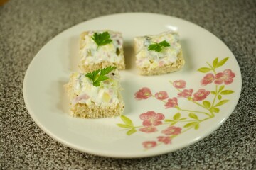 Three creamy mini sandwiches garnished with fresh parsley leaves, served on a floral plate against a textured surface for a delightful presentation.