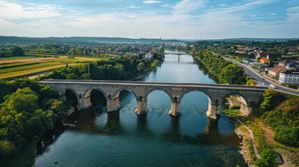 Naklejka premium Arched bridge crosses river through green landscape under blue sky.