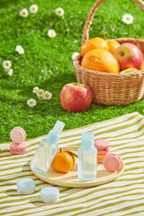 High angle shot on a picnic carpet, which spread on green grass. Some unlabeled skincare bottles displayed on a round dish with small cakes and fresh fruits