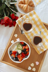Breakfast tray with coffee, fruit bowl, croissants, and roses on bed.