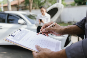 Asian woman and insurance agent assess damaged car after accident on roadside. One uses smartphone for communication, emphasizing concern and professionalism in handling claims and repair process.