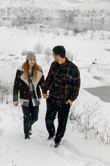 a couple walking along the snowy shore of a pond. A man and a woman are holding hands and smiling at each other. Both are dressed in winter clothes